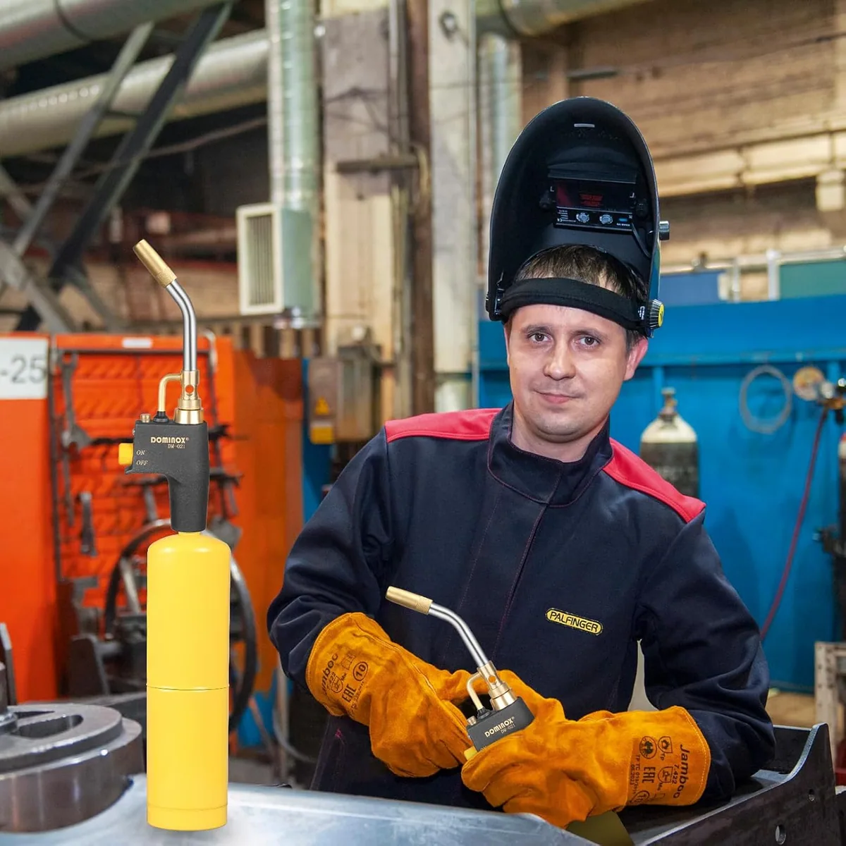 A man in a welding mask and gloves holding a DOMINOX torch in a metal shop
